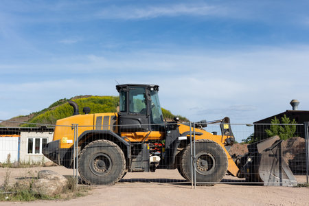 Bright yellow wheel loader stands behind metal fence on dusty construction ground. Sunlight...