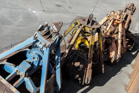 Row of heavy grab buckets rests on concrete surface, showing worn paint, rust textures and complex metal mechanics. Industrial tools create rugged atmosphere with strong sunlight and sharp shadowsの写真素材