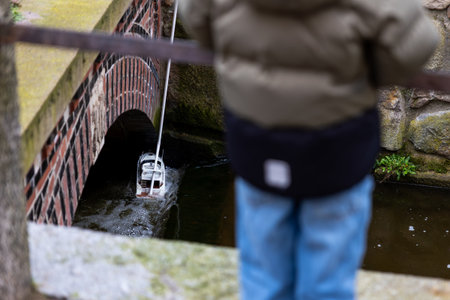 Child controls remote model boat navigating narrow canal under brick bridge visible water current reflections. Urban outdoor leisure scene shows childhood hobby miniature technologyの写真素材