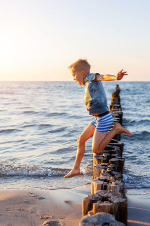Happy child jumping barefoot from wooden groyne shallow sea during warm sunset, showing freedom courage energy playful summer lifestyle. Golden sunlight beach waves motion, outdoor childhood momentの写真素材