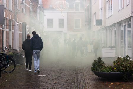Football fans crowd fills narrow street green smoke bomb thick haze at brick facades glowing lights. Silhouettes movement passion shape intense urban atmosphere celebration in Deventer Netherlandsの写真素材
