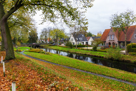 Hattem town Netherlands canal brick houses autumn foliage scenic idyllic landscape view. Quiet residential neighborhood with trees reflections pathway under cloudy sky peaceful travel atmosphereの写真素材