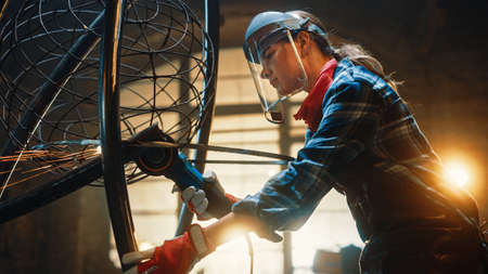 Young Contemporary Female Artist is Grinding Her Metal Tube Sculpture with a Handheld Power Tool in a Studio Workshop. Empowering Woman Makes Modern Brutal Abstract Artwork Out Of Metal.の写真素材