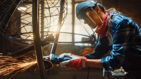 Close Up of Young Female Fabricator in Safety Mask. She is Grinding a Metal Tube Sculpture with an Angle Grinder in a Studio Workshop. Empowering Woman Makes Modern Abstract Metal Artwork.の写真素材