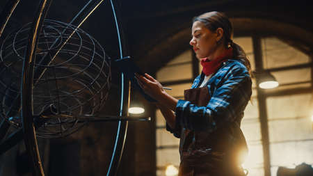 Beautiful Female Artist Sketches on a Tablet Computer Next to Brutal Metal Sculpture in Studio....