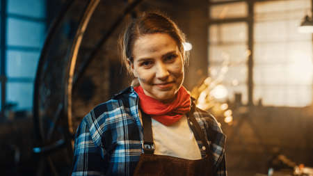 Young Beautiful Empowering Woman with Ear Piercing Gently Smiles at the Camera. Authentic Fabricator Wearing Work Clothes in a Metal Workshop. Sparks Flying on the Background.の写真素材