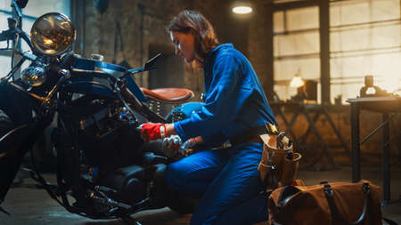 Young Beautiful Female Mechanic is Working on a Custom Bobber Motorcycle. Talented Girl Wearing a Blue Jumpsuit. She Uses a Ratchet Spanner to Tighten Nut Bolts. Creative Authentic Workshop Garage.の写真素材