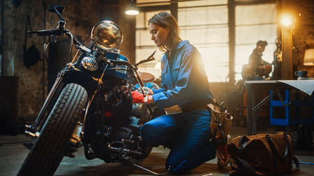 Young Beautiful Female Mechanic is Working on a Custom Bobber Motorcycle. Talented Girl Wearing a Blue Jumpsuit. She Uses a Ratchet Spanner to Tighten Nut Bolts. Creative Authentic Workshop Garage.の写真素材