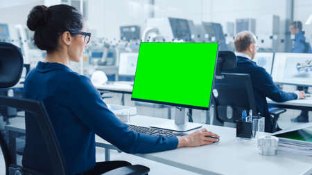 Over the Shoulder: Female Chief Engineer Working on Personal Computer, Display is Green Mock-up Screen or Chroma Key. Industrial Factory with High-Tech Machinery and Professional Workersの写真素材