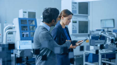 Modern Factory: Female Project Manager and Male Engineer Walk Through High Tech Development Facility, Talking and Using Tablet Computer. High-Tech Facility with CNC Machinery Robot Arm Production Lineの写真素材
