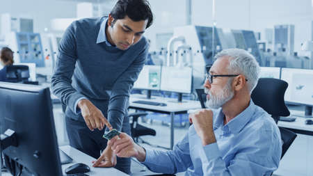 Modern Factory Office: Indian Project Manager Talks to Caucasian Senior Engineer who Working on...
