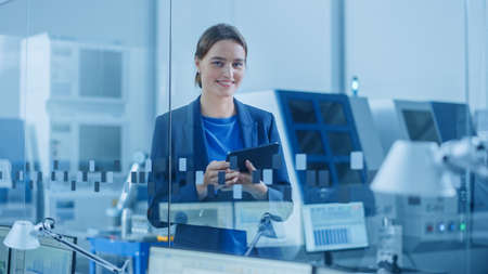 Modern Factory Office: Portrait of Young and Confident Female Industrial Engineer Standing and Holding Digital Tablet. Industrial Factory Office with Glass Wall and CNC Machinery Workshop Behindの写真素材