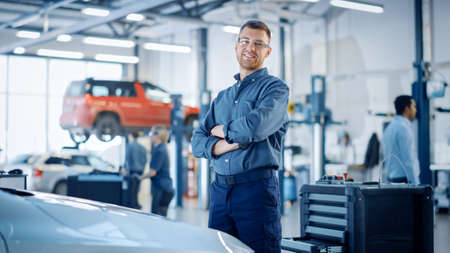 Handsome Car Mechanic is Posing in a Car Service. He Wears a Jeans Shirt and Safety Glasses. His Arms are Crossed. Specialist Looks at a Camera and Smiles.の写真素材