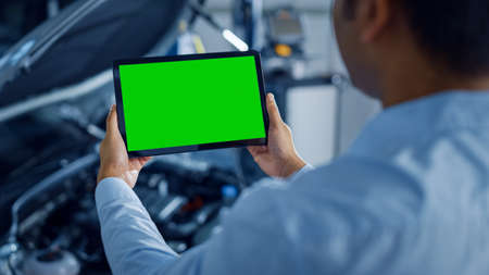 Car Service Manager or Mechanic Uses a Tablet Computer with a Green Screen Mock Up that is Pointed at an Enginer Bay. Specialist Inspecting the Vehicle in Order to Find Broken Components In the Engineの写真素材