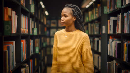 Portrait of Gifted Beautiful Black Girl Stands Between Rows of Bookshelves Searching for the Right Book Title for Class Assignmentの写真素材