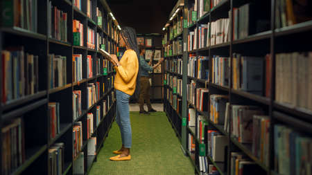 University Library: Shot of Gifted Beautiful Black Girl Stands Between Rows of Bookshelves Searching for the Right Book for Class Assignment. Focused Smart Student Learning, Studying for Examsの写真素材