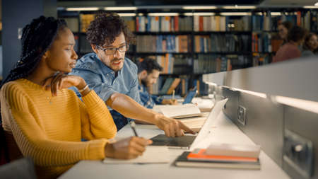 University Library: Gifted Black Girl uses Laptop, Smart Classmate Explains and Helps Her with Class Assignment. Study for Class Writing Paper Assignment. Diverse Students Learning, Studying for Examsの写真素材
