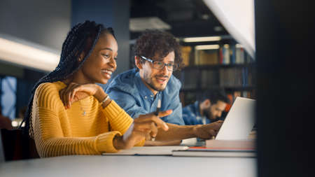 University Library: Gifted Black Girl uses Laptop, Smart Classmate Explains and Helps Her with Class Assignment. Happy Diverse Students Talking, Learning, Studying Together for Examsの写真素材