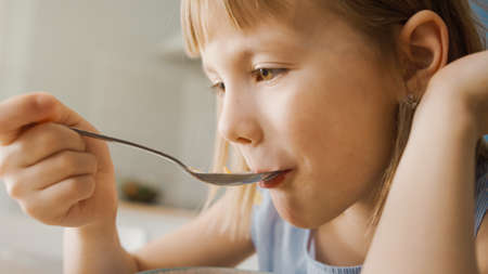 Breakfast in the Kitchen: Portrait of Adorable Little Girl Eating Healthy Granola Cereal with Milk out of Bowl. Cute Little Girl having Breakfast. In the Background Caring Mother Prepares Foodの写真素材