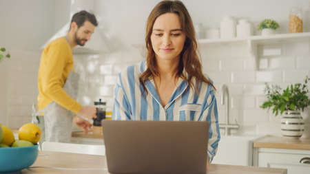 Young Beautiful Woman Using Laptop Computer While Wearing Blue Pyjamas. Brunette Female Sitting in a Modern Sunny Kitchen Room. Freelancer Working from Home.の写真素材