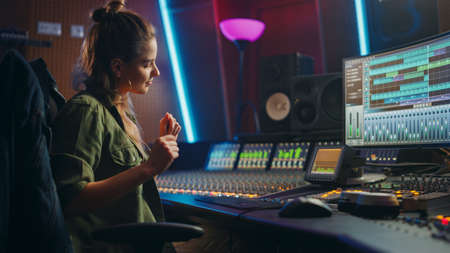 Stylish Female Audio Engineer Working in Music Recording Studio, Uses Mixing Board, Software to Create Modern Sound. Creative Girl Artist Musician Working on Control Desk to Produce New Song.の写真素材