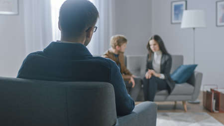 Unfocused Couple on Counseling Session with Psychotherapist. Focus on Back of Therapist Taking Notes and Talking: People Sitting on Analyst Couch, Discussing Psychological and Relationship Problemsの写真素材