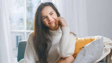 Shot of Beautiful Young Brunette Smiling Charmingly while Looking at Camera. Shy Girl Wearing White Knitted Sweater Laughing in Cozy Roomの写真素材