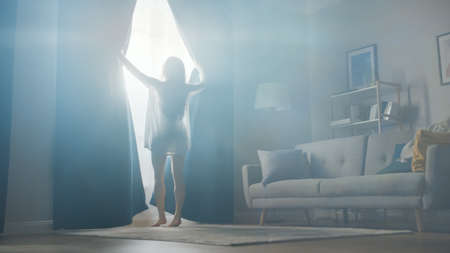 Beautiful Young Brunette Opens Curtains To let Morning Sunlight Into Cozy Living Room. Sensual Girl Rising Early and Greeting New Day.の写真素材