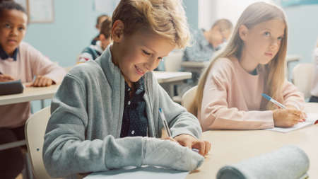 In Elementary School Classroom Brilliant Caucasian Boy Writes in Exercise Notebook, His Hand in a...