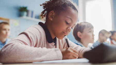 In Elementary School Classroom Black Girl Writes in Exercise Notebook, Taking Test. Junior Classroom with Diverse Group of Bright Children Working Diligently, Learning. Low Angle Side View Portraitの写真素材