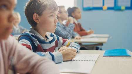 In Elementary School Class: Portrait of Brilliant Caucasian Boy Writes in Exercise Notebook, Taking Test and Writing Exam. Diverse Group of Bright Children Working Diligently and Learningの写真素材