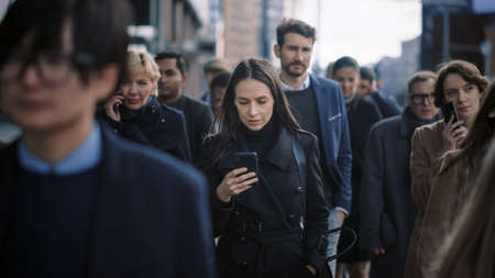 Beautiful Businesswoman in Black Coat is Using a Smartphone on a Street in Downtown. She Walks on a Crowded Pedestrian Street and Looks Successful. Shes Browsing the Web on Her Device.の写真素材