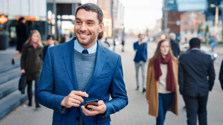 Caucasian Businessman in a Suit is Using a Smartphone on a Street in Downtown. Other Office People Walk Past. He Smiles and Looks Successful. Hes Browsing Web on his Device. Shot with Warm Sun Flare.の写真素材