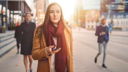 Young Smart Casually Dressed Female is Using a Smartphone on a Street. Business People and Office Managers Walk Pass on Their Way to Work. She Looks Confident while Checking Her Cell and Walking.の写真素材