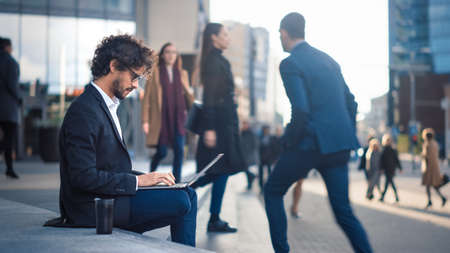 Handsome Businessman in a Suit is Sitting on Steps next to Business Center and Working on a Laptop on a Street in a City. Office People Walk By to Work.の写真素材