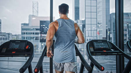 Athletic Muscular Man Running on a Treadmill, Leg and Cardio Day. Strong Man Training in the Modern Gym Fitness Club with Window Cityscape View. Back View Shotの写真素材