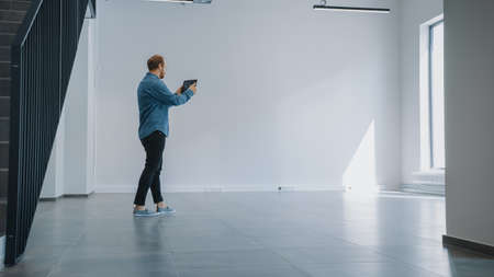 Young Hipster Man in Jeans Shirt Standing in Empty Office and Map it with an Augmented Reality Software on a Tablet. Sunlight Shines Through Big Windows.の写真素材
