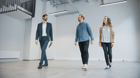 Real Estate Agent Showing a New Empty Office Space to Young Male and Female Hipsters. Entrepreneurs Meet the Broker with a Tablet and Discuss the Facility They Wish to Purchase or Rent.の写真素材