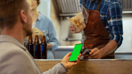 Food Truck Employee Hands Out a Freshly Made Burger to a Happy Young Male. Man in a Casual Suit is Using His Smartphone with Green Screen Mock-Up Template for Mobile Payment Solution.の写真素材