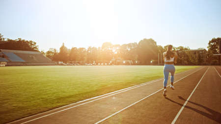 Beautiful Fitness Girl in Light Blue Athletic Top and Leggings Jogging in the Stadium. She is Running on a Warm Summer Afternoon. Athlete Doing Her Routine Sports Practice on a Track.の写真素材