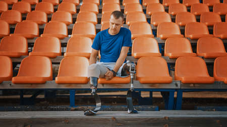 Athletic Disabled Fit Man with Prosthetic Running Blades is Preparing for a Training on an Outdoor Stadium on a Sunny Afternoon. Amputee Runner is Sitting and Fixates His Legs for a Run.の写真素材