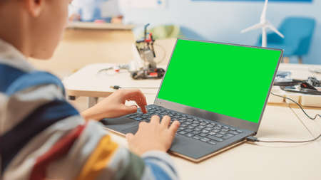 Elementary School Computer Science Classroom: Over the Shoulder View of a Kid Using Green Chroma Key Screen Laptop to Program Software for Robotics Engineering Classの写真素材