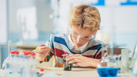 Elementary School Science or Chemistry Classroom: Smart Little Boy wearing Safety Glasses Mixes Chemicals in Beakersの写真素材