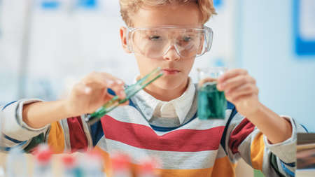 Elementary School Science or Chemistry Classroom: Smart Little Boy wearing Safety Glasses Mixes Chemicals in Beakersの写真素材