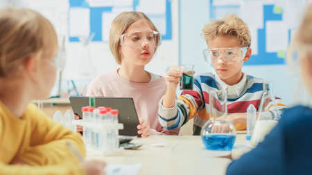 Elementary School Science Classroom: Smart Boy Mixes Chemicals in Beakers and Cute Girl Writes Down and Analyzes Results with Digital Tablet Computer. Modern Education, Learning Chemistryの写真素材