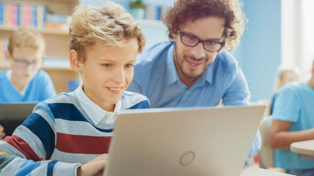 Elementary School Computer Science Class: Smart Boy Uses Laptop Computer, Friendly Teacher Helps Him...
