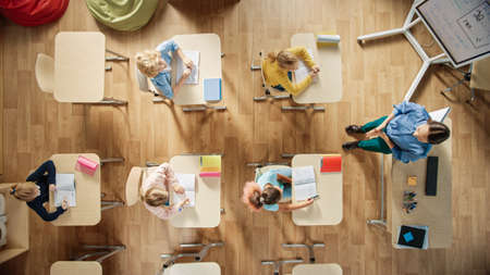 Bright Elementary School Classroom: Children Sitting at their School Desk Working, Doing Assignment, Enthusiastic Teacher Stands a the Head of the Class Explaining a Lesson. Top View Shot.の写真素材