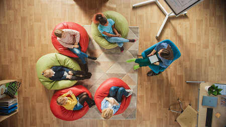 Elementary School Creativity Classroom: Children Sitting on the Bean bags while Teacher Explains Lesson to them and Uses Digital Tablet Computer. Top View Shotの写真素材