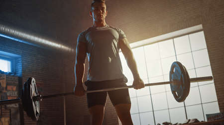 Handsome Muscular Man Does Overhead Deadlift with a Barbell in a Small Authentic Gym. Athletic Man Training His Arm Muscles and Exercises with Barbell. Workout in the Hardcore Gym. Warm Light.の写真素材