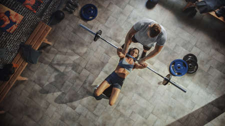 Top View of Professional Female Bodybuilder Doing Bench Press Workout with a Barbell in the Hardcore Gym, Trainer Partner Assists with Equipment and Trauma Prevention. Couple Doing Barbell Exerciseの写真素材